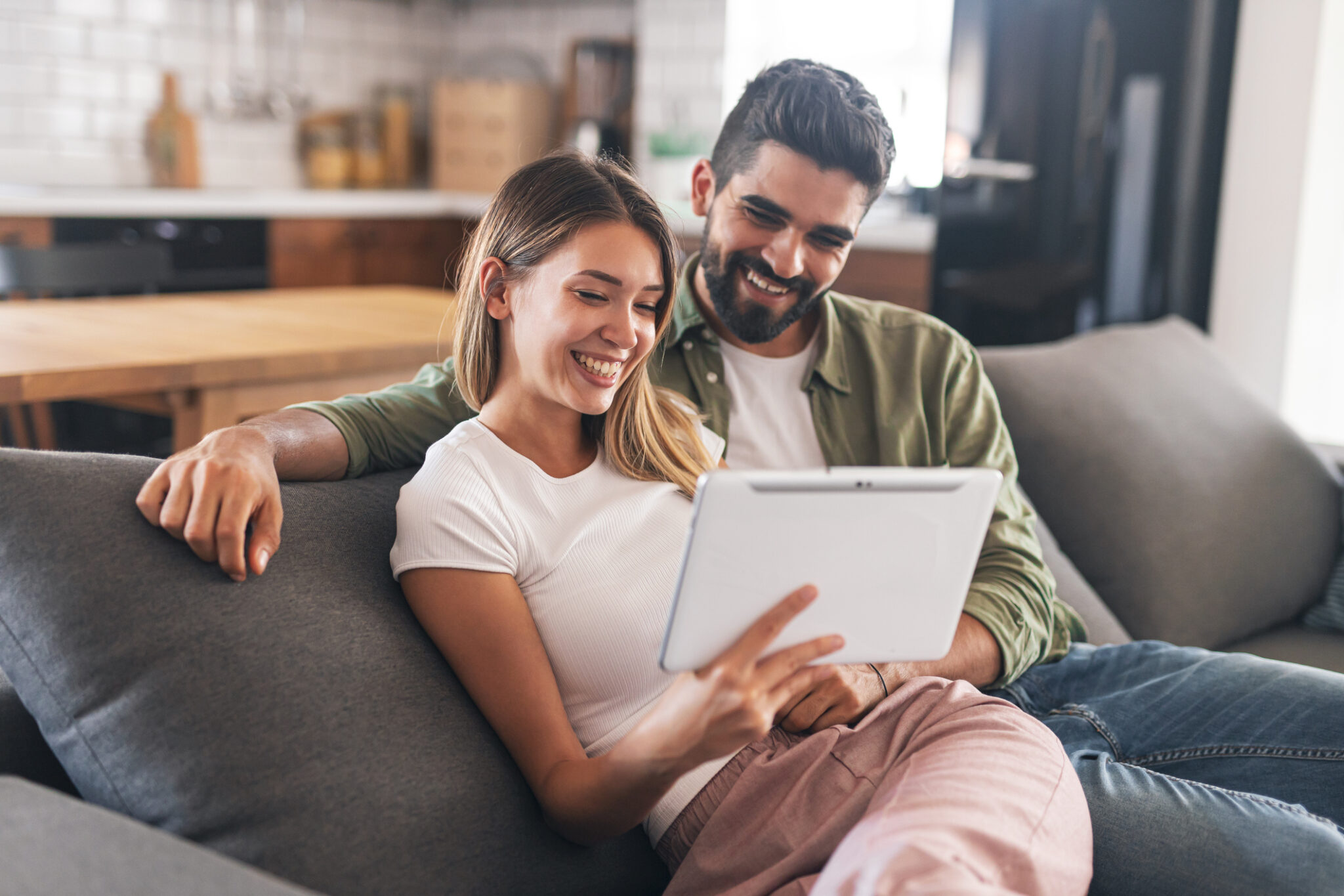 A smiling couple relaxing on their living-room sofa while reading customer reviews on a tablet, reflecting the positive experiences of homeowners who use the MyPlaceIQ smart home system every day.