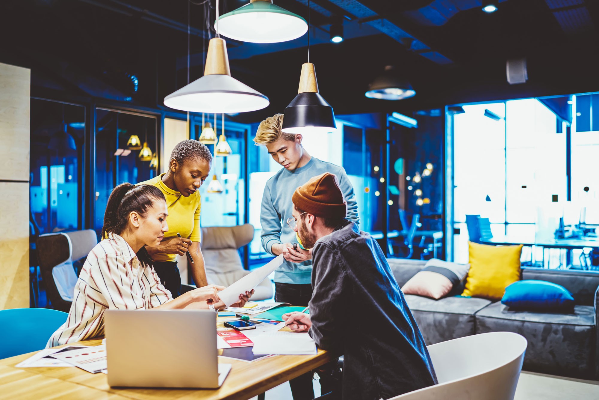 A diverse group of young professionals collaborating around a table in a modern, creative workspace with vibrant lighting and comfortable seating.