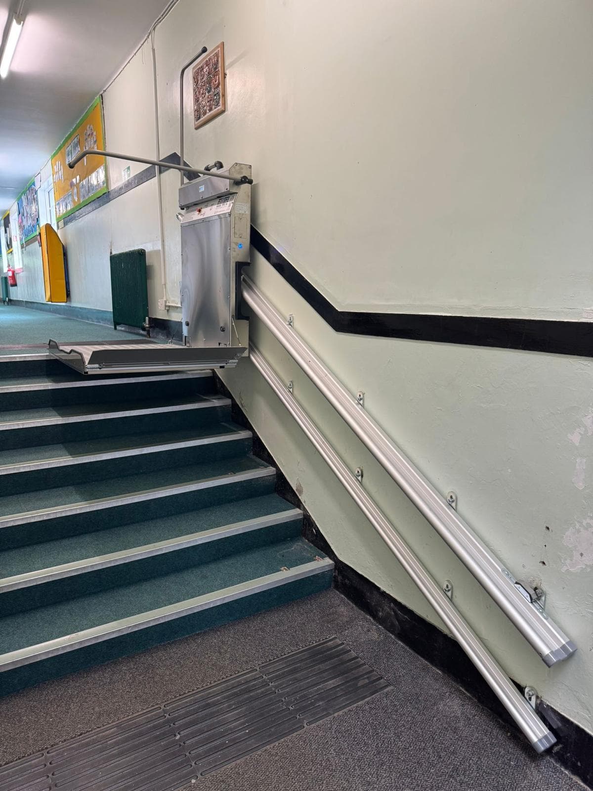 Commercial inclined platform stairlift in folded position alongside carpeted internal stairs in a UK school or public building — wall-mounted rail with aluminium chassis and folded platform at lower landing