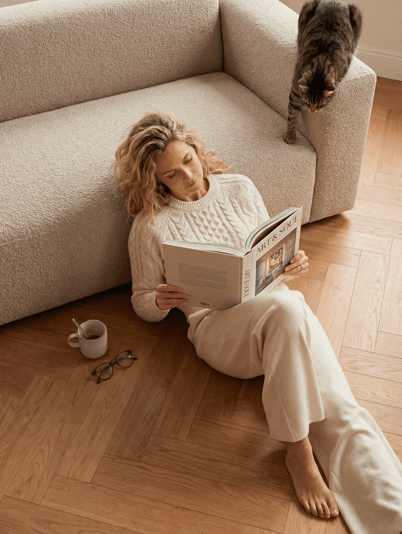 Woman with curly hair sitting on the floor against a cream sofa reading a magazine, with a tabby cat climbing over the sofa behind her.
