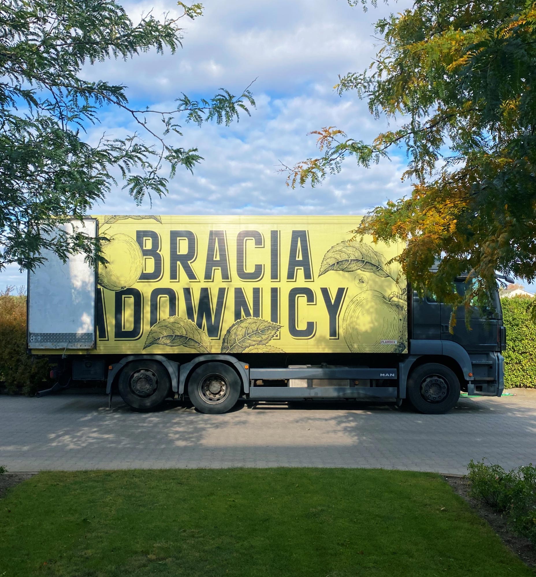 Ciężarówka z żółtozieloną naczepą stoi przy drzewach; na boku wielki napis „BRACIA SADOWNICY” i szkice jabłek.