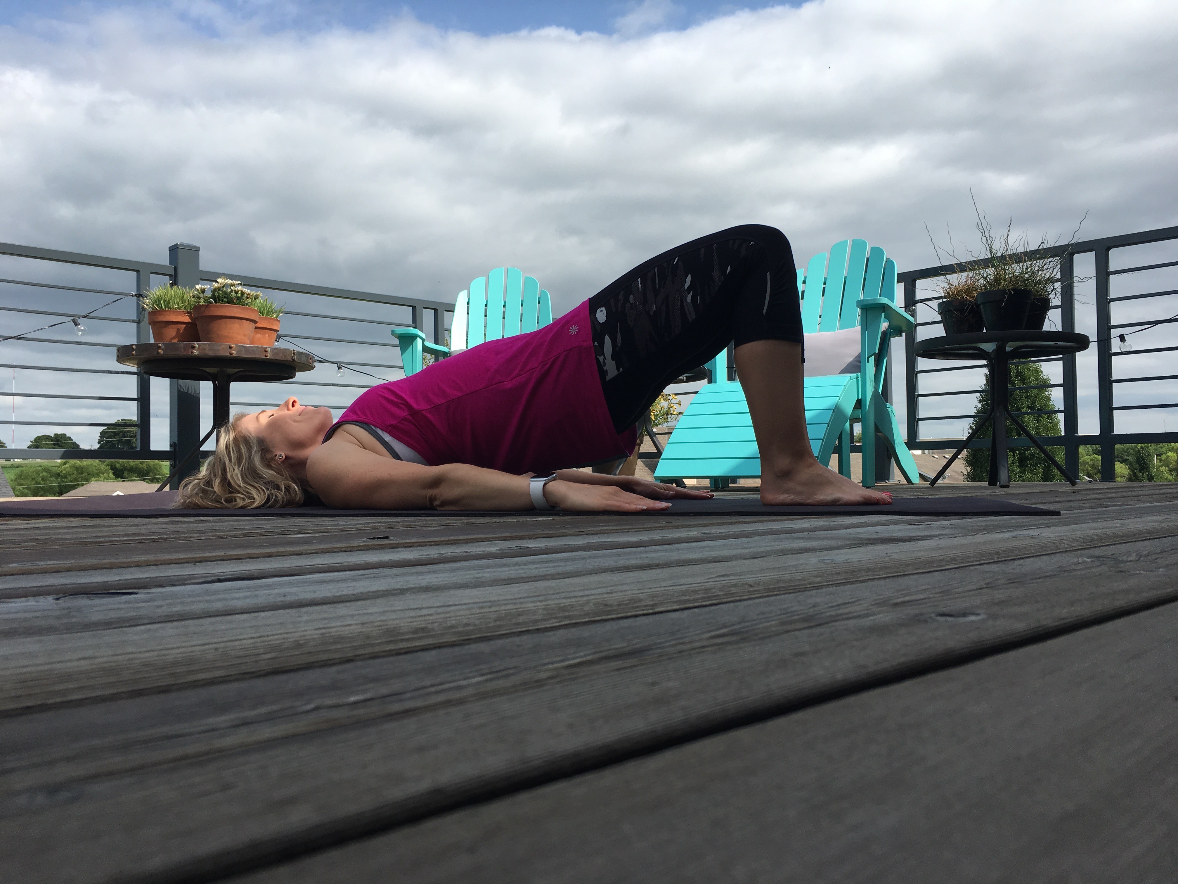 Heather Hausman is practicing yoga outdoors on a wooden deck, performing a Superman pose with overcast skies in the background, surrounded by blue chairs and potted plants.