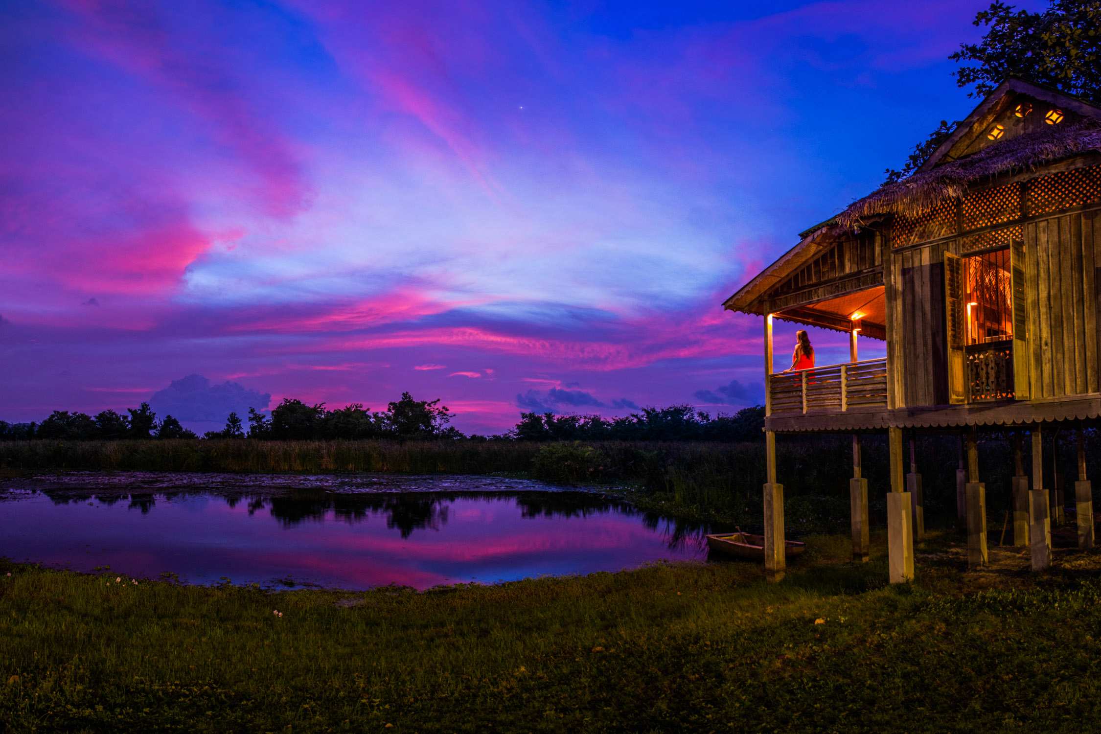A wooden stilt house overlooks a lake under a vibrant pink and blue sunset sky with wispy clouds.