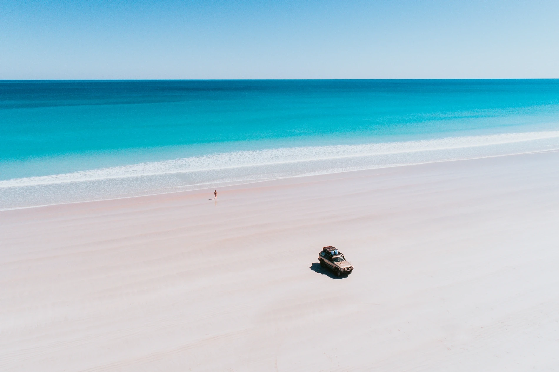 A vast empty beach with a four-wheel drive parked on the sand and turquoise surf beyond