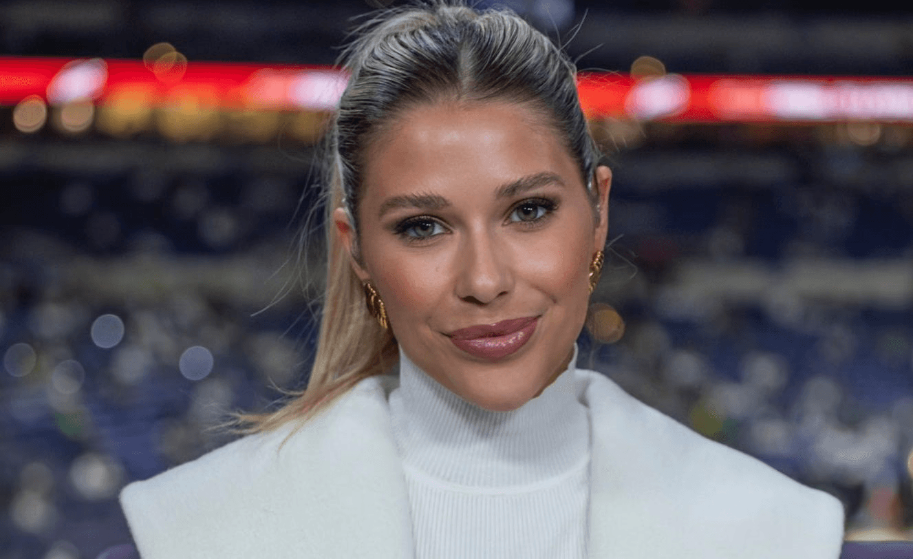 Jordan Giorgio with hair tied back, wearing a white turtleneck and white coat, stands indoors with blurred stadium seats and lights in the background, smiling at the camera.