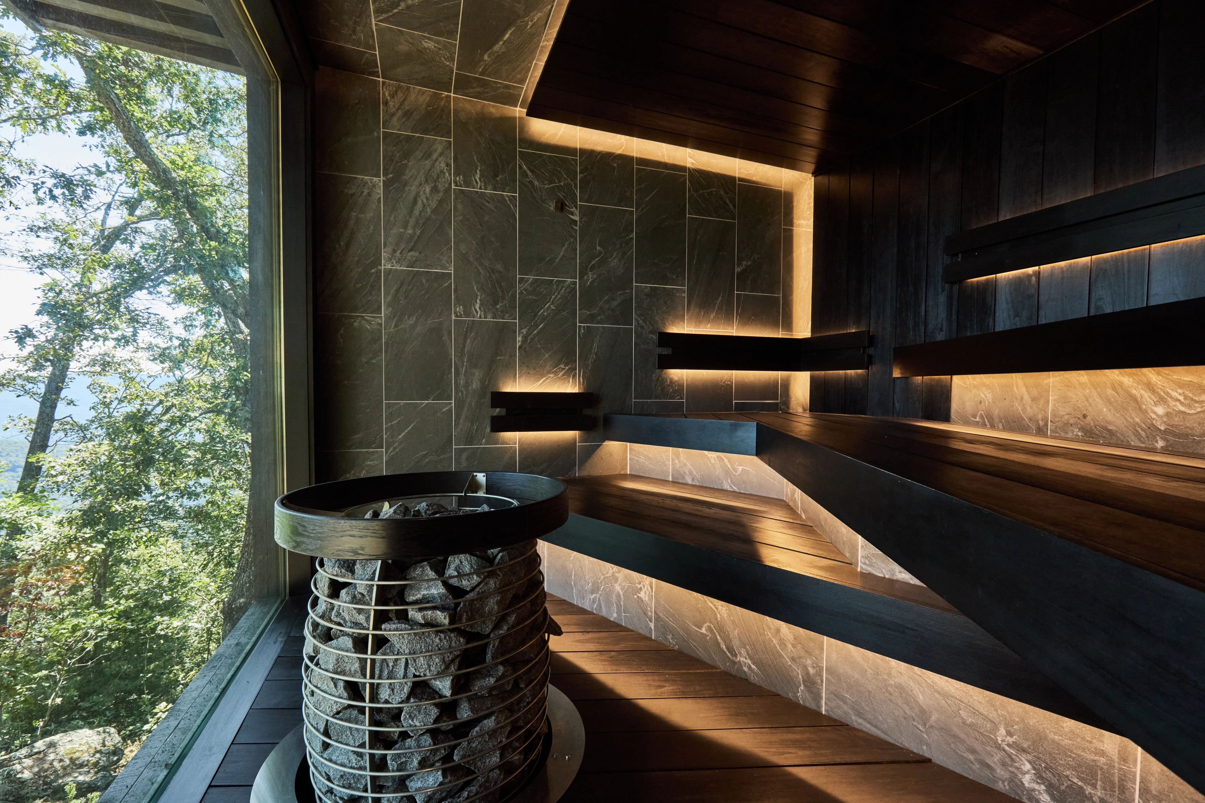 Sauna interior with dark wood benches, warm concealed lighting, a stone-filled heater and a full-height window overlooking trees.