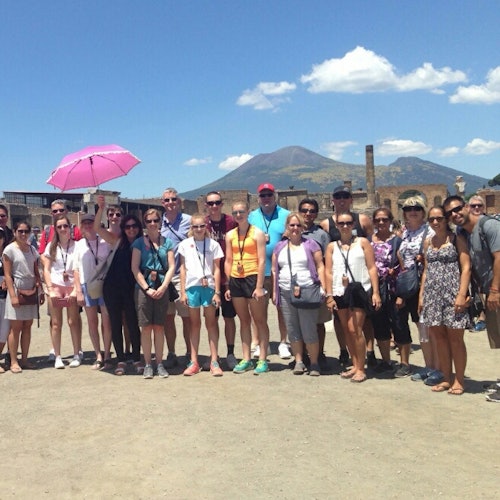 A group of people standing outdoors with ruins and a mountain in the background. One person holds a pink umbrella.