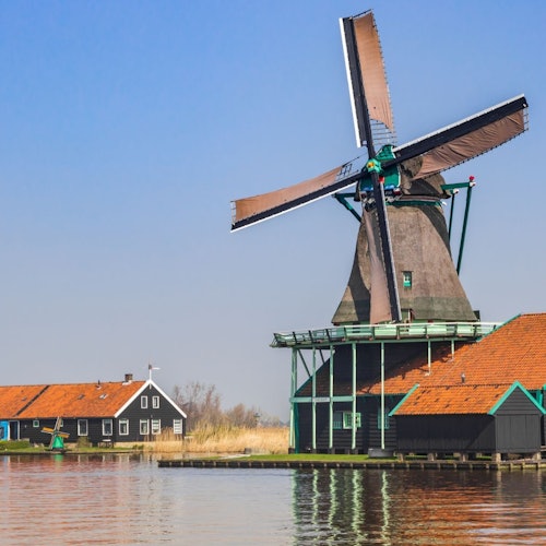 A windmill with large blades is situated next to a building with a red roof by a calm waterway on a clear day.