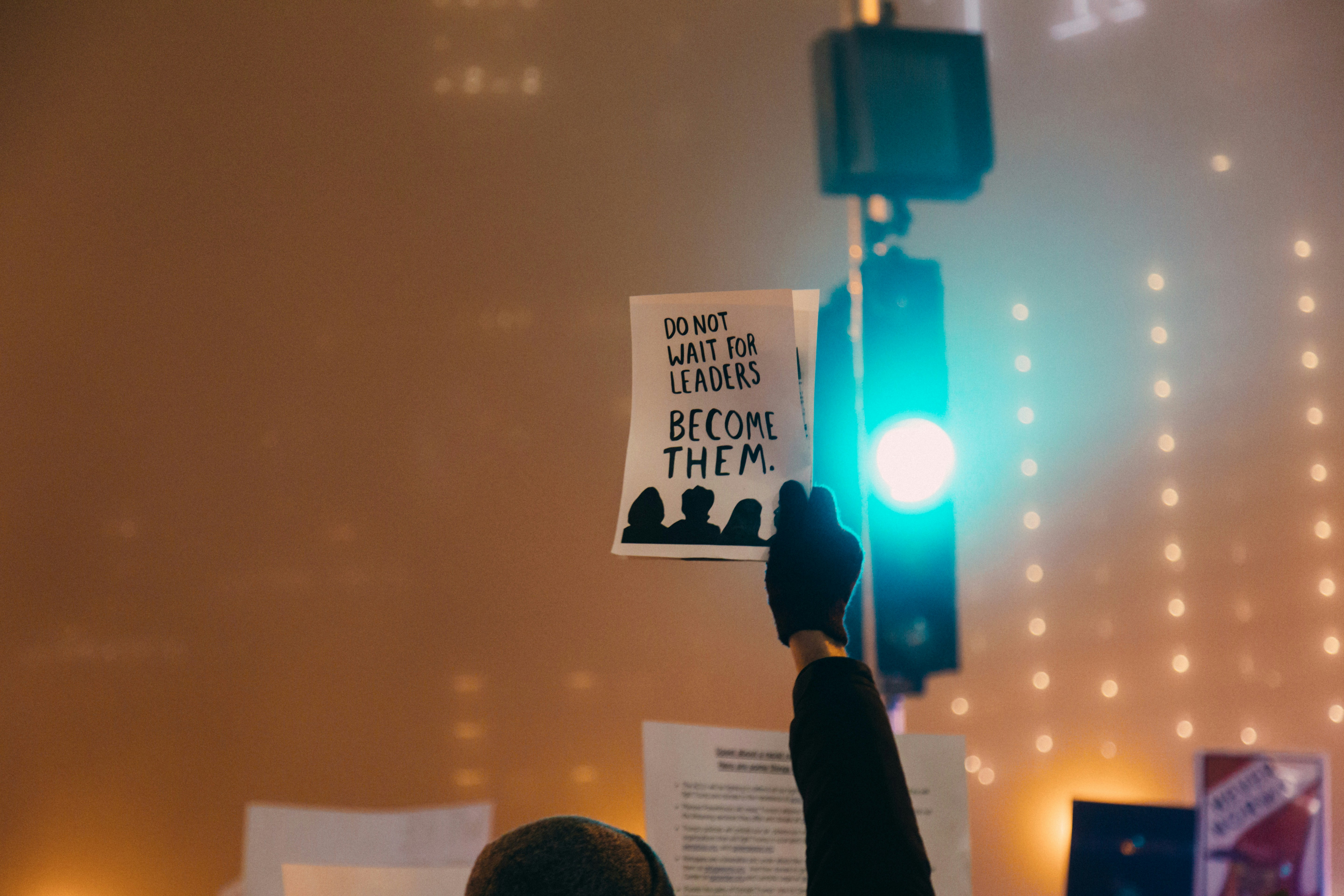 Personne tenant une pancarte avec le message 'Do not wait for leaders, become them' lors d'une manifestation, avec des lumières en arrière-plan