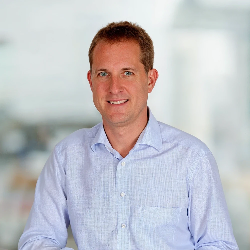 A smiling individual in a light blue shirt stands in a bright, blurred indoor setting.