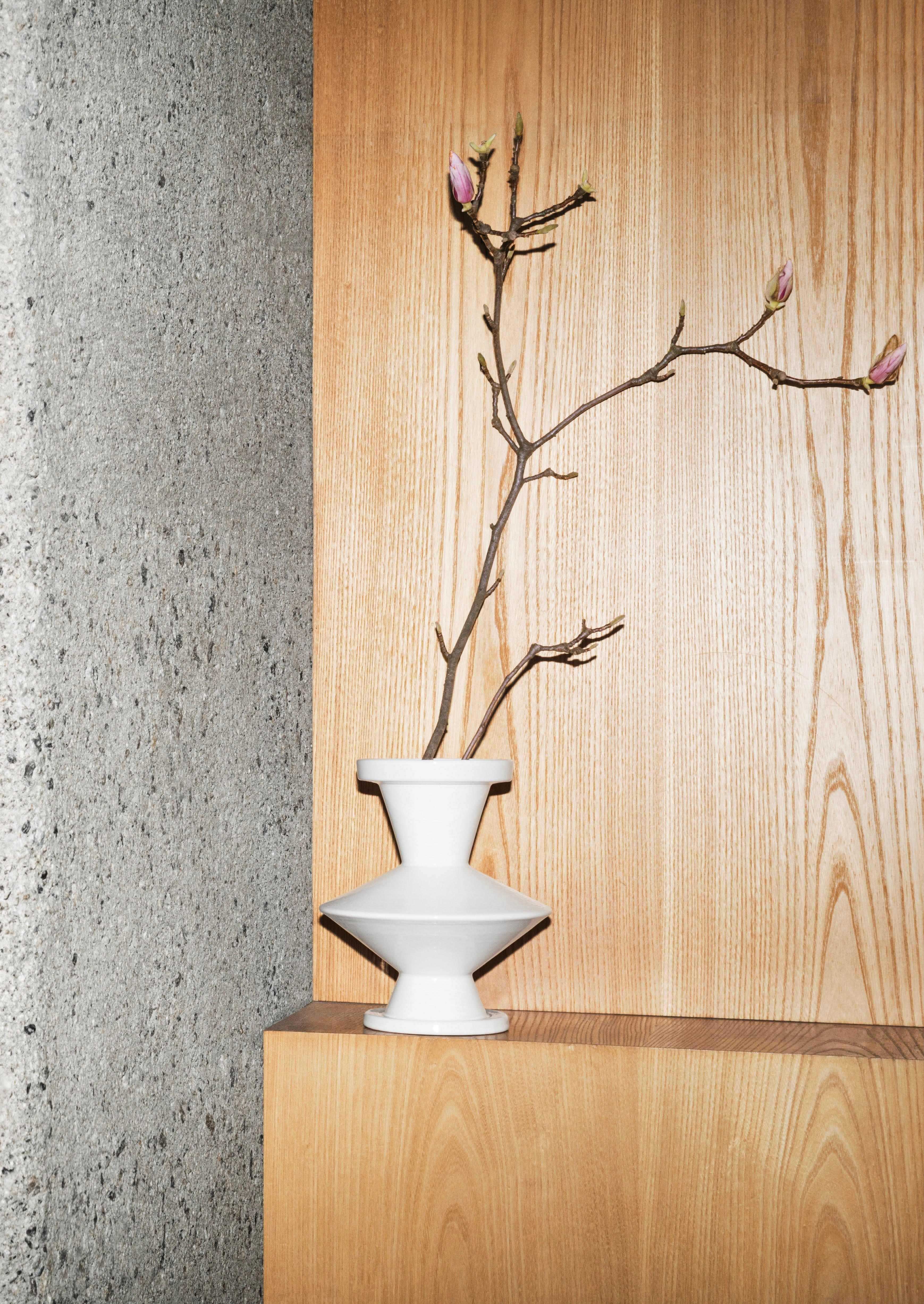 A close-up shot of architectural details. On the left are a series of vertical copper panels. In the center is a textured concrete column. On the right, a simple white vase with a flowering branch is placed on a wooden shelf against a wooden wall.
