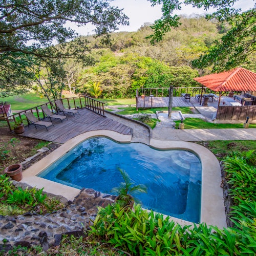 สระว่ายน้ำในร่มล้อมรอบด้วยพื้นที่สีเขียว พื้นไม้พร้อมเก้าอี้นั่งเล่น และศาลาพร้อมหลังคาสีแดงในพื้นที่ป่าที่เขียวขจี