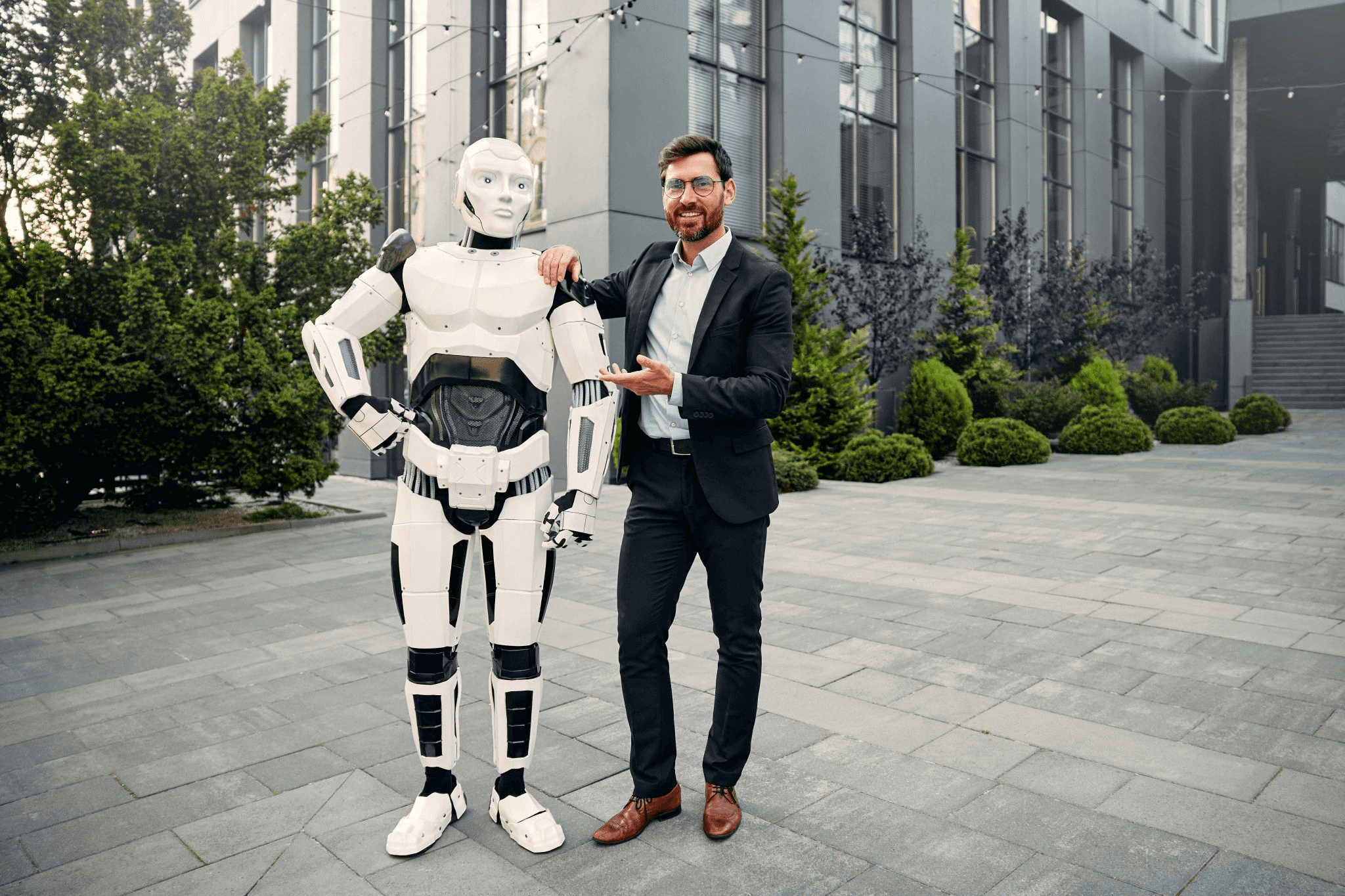 A man in a suit standing beside a white humanoid robot outside a modern office building.
