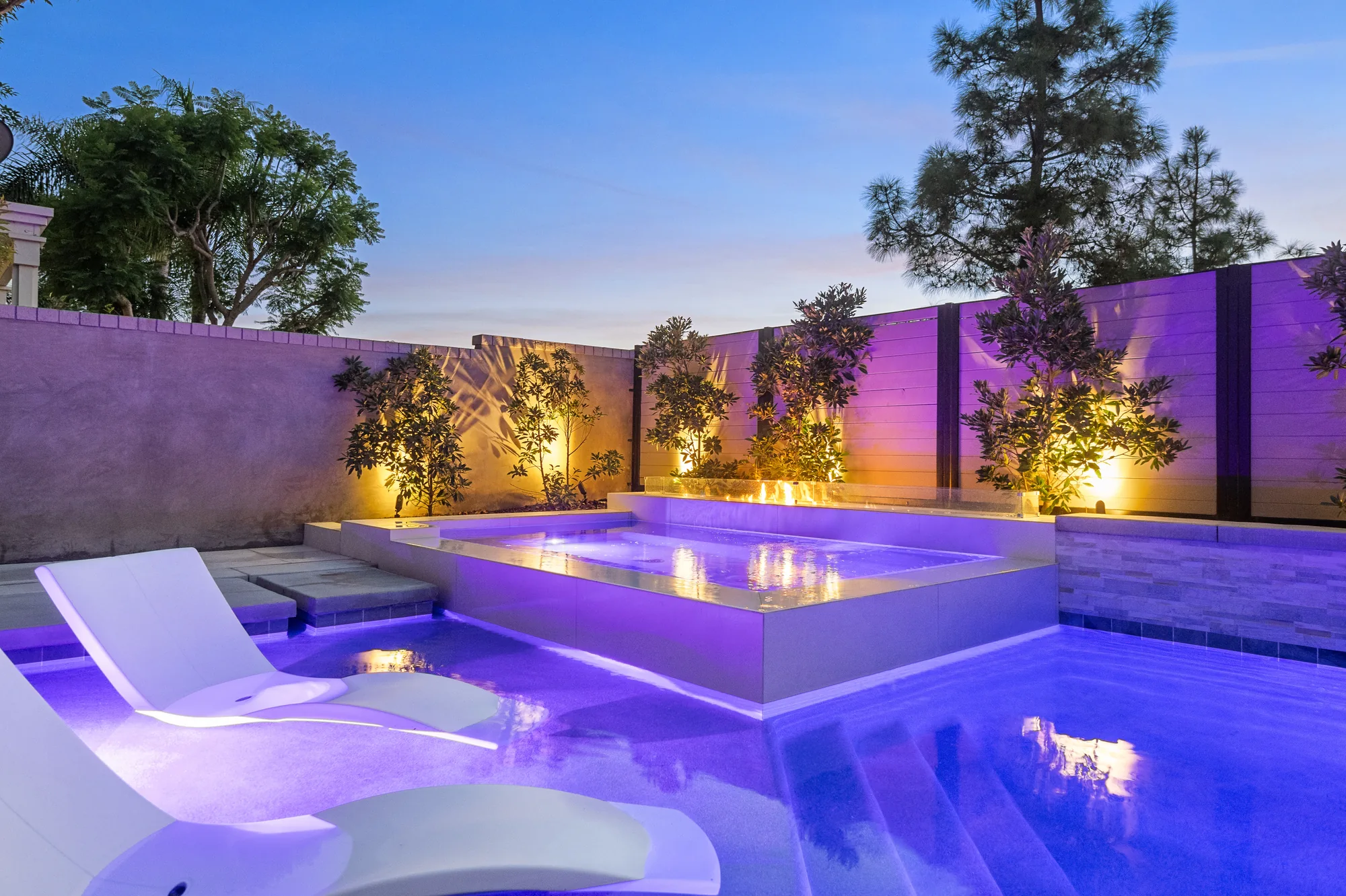 Illuminated raised spa and white loungers set within a pool beside planted boundary walls.