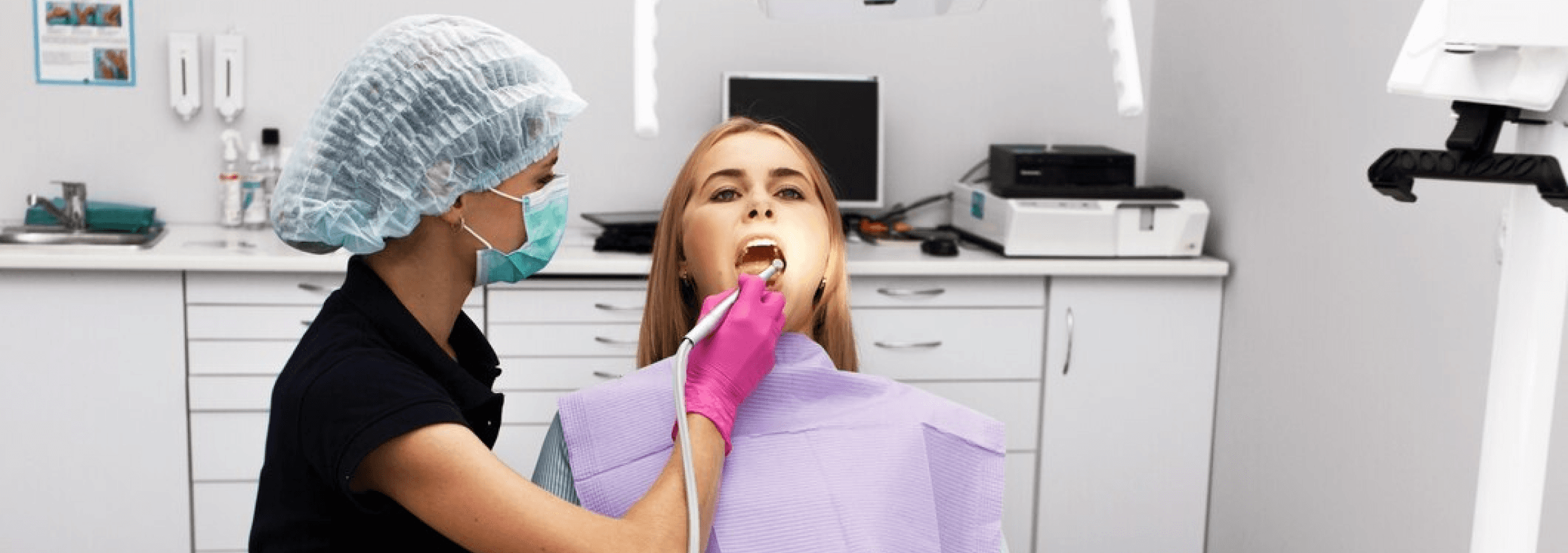 A dentist wearing a mask and hair covering performing a dental procedure on a patient in a dental chair, using a dental instrument while the patient wears a protective bib.