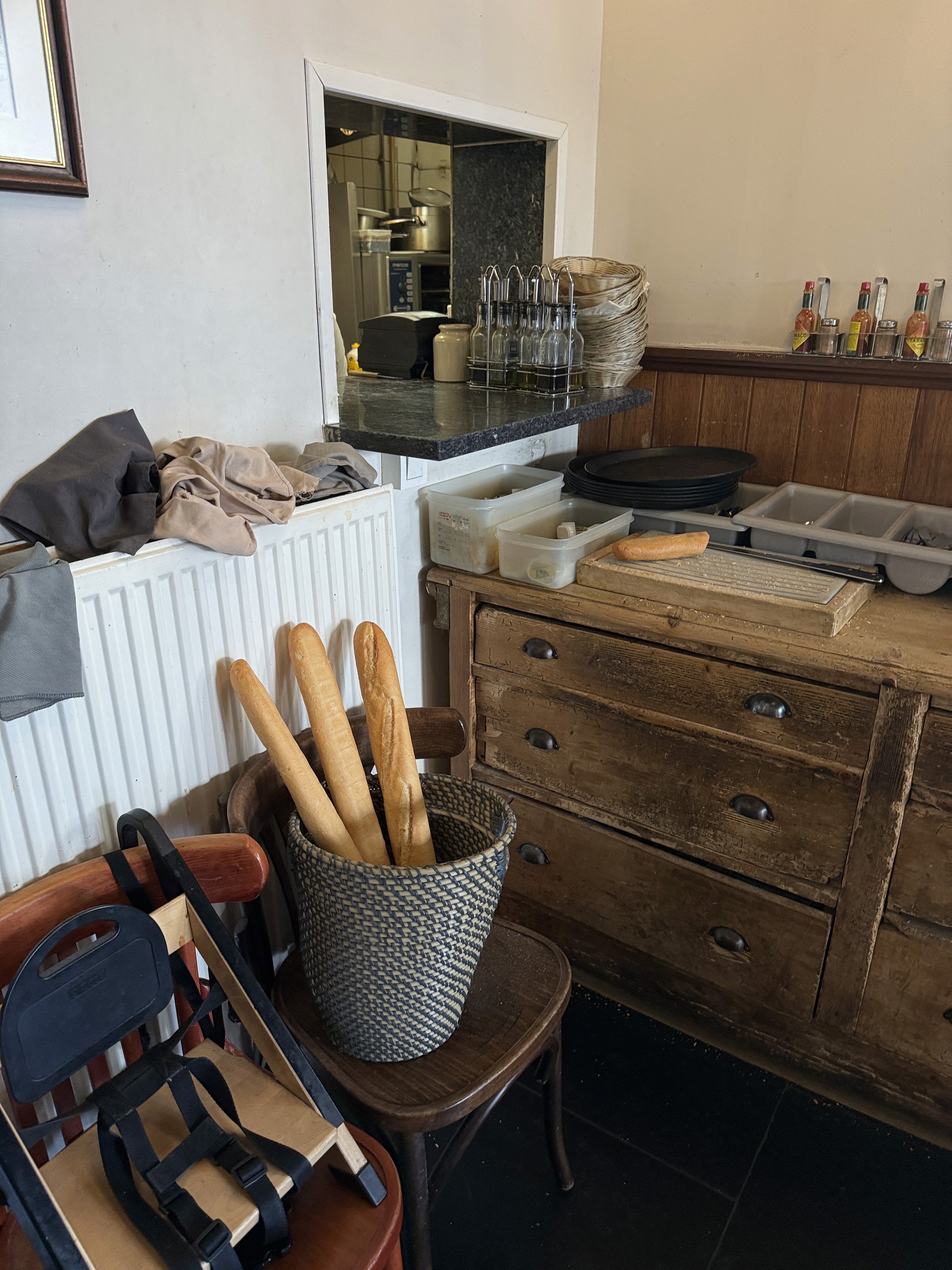 Três baguetes em um cesto de palha trançada, colocadas sobre uma cadeira de madeira em um ambiente de cozinha rústica. À direita, um balcão de madeira antiga com gavetas e, sobre ele, uma bancada preta com tábuas de corte, recipientes de plástico e garrafas com temperos ao fundo.