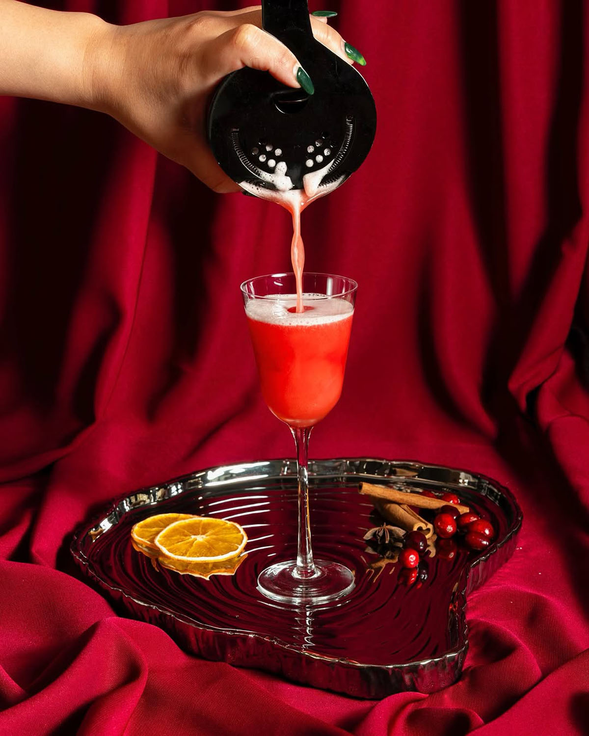 A vibrant red cocktail being poured into an elegant glass at Node Studio, styled on a heart-shaped glass tray with citrus and cranberries, set against a dramatic crimson backdrop