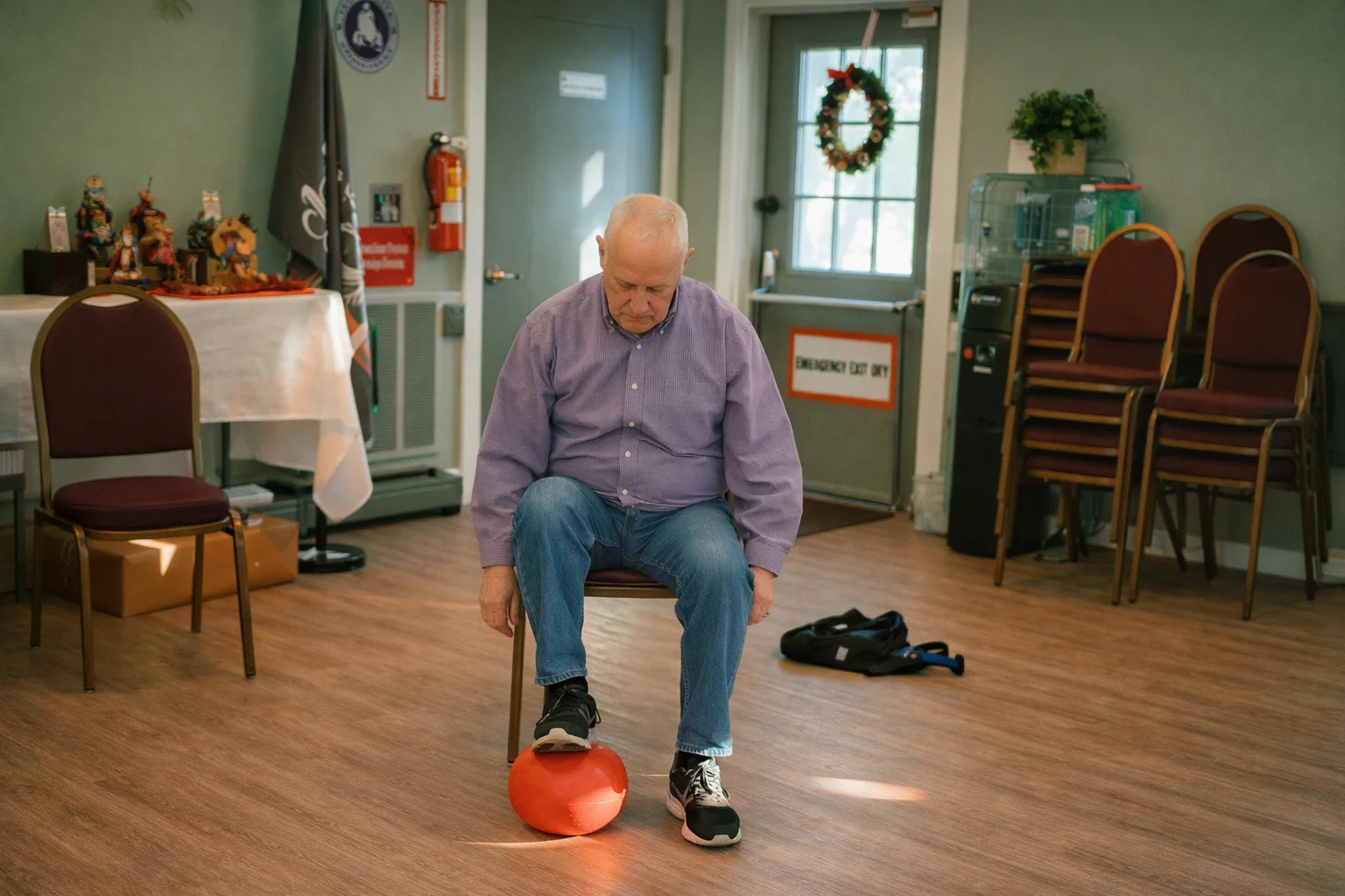 An elderly man sits on a chair, focusing on an orange object on the floor in a room with chairs and decor.