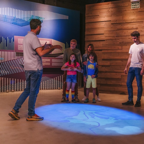 Eine Gruppe von Menschen steht in einem Ausstellungsraum mit blauen und holzgetäfelten Wänden, und es gibt eine Projektion auf dem Boden.