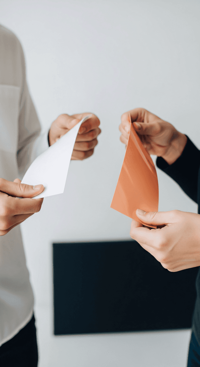 Two people each holding a different sheet of paper, one white and one orange, suggesting a comparison or handoff between two versions
