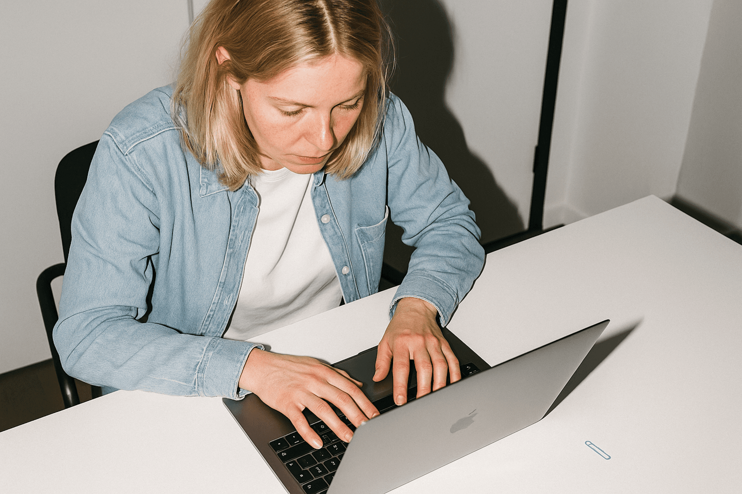 Woman with blonde hair typing on a laptop at a white table, focused on her work. Soft lighting in the background.