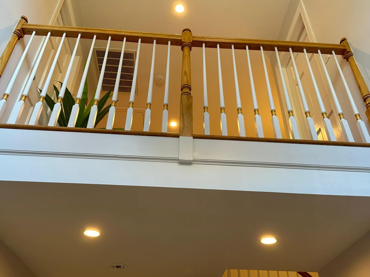 Second-floor balcony with white wooden balusters featuring gold accents and a natural wood handrail, viewed from below under warm recessed lighting.