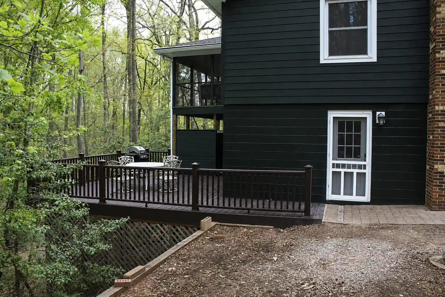 Exterior view of a screened-in porch above a wood deck, surrounded by mature trees, with outdoor dining furniture and a grill.
