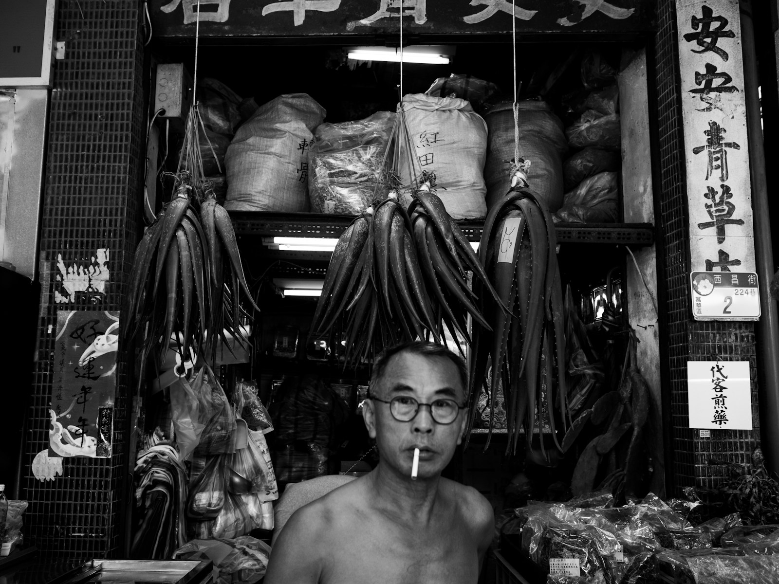 A stallholder smoking beneath hanging produce at a night market