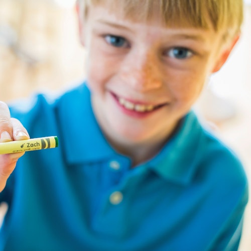 微笑的孩子穿著藍色衫，手持一支標有「Zach」的黃綠色蠟筆，靠近鏡頭。