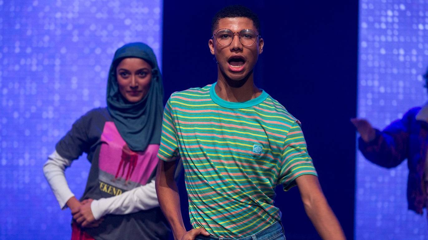 A young non-binary performer in a striped shirt sings expressively downstage, with a performer in a hijab standing behind him under purple stage light.