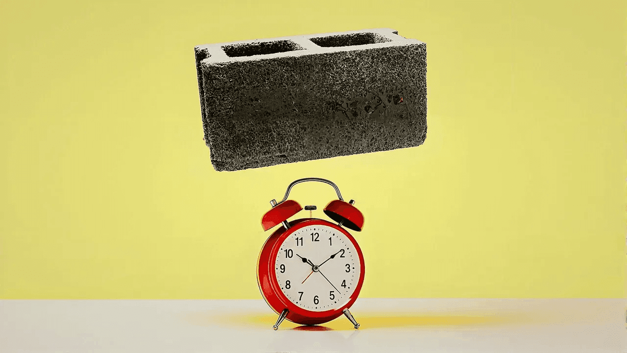 Cinder block about to fall on red alarm clock on white surface, yellow background