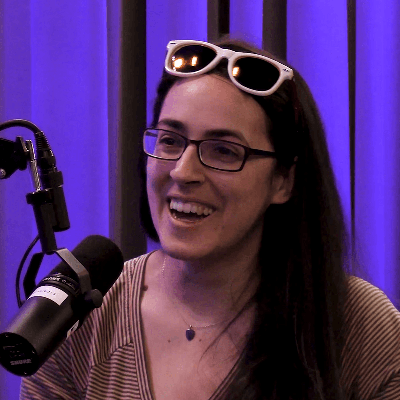 A podcast recording scene featuring a smiling individual in glasses with white sunglasses resting on their head, speaking into a studio microphone. The background is softly lit with purple curtains, giving the setup a professional and creative atmosphere.