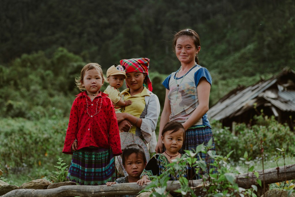 A family of five, including three young children, a mother, and an older girl, stands in a lush, rural setting with traditional attire, highlighting their cultural heritage and connection to nature.