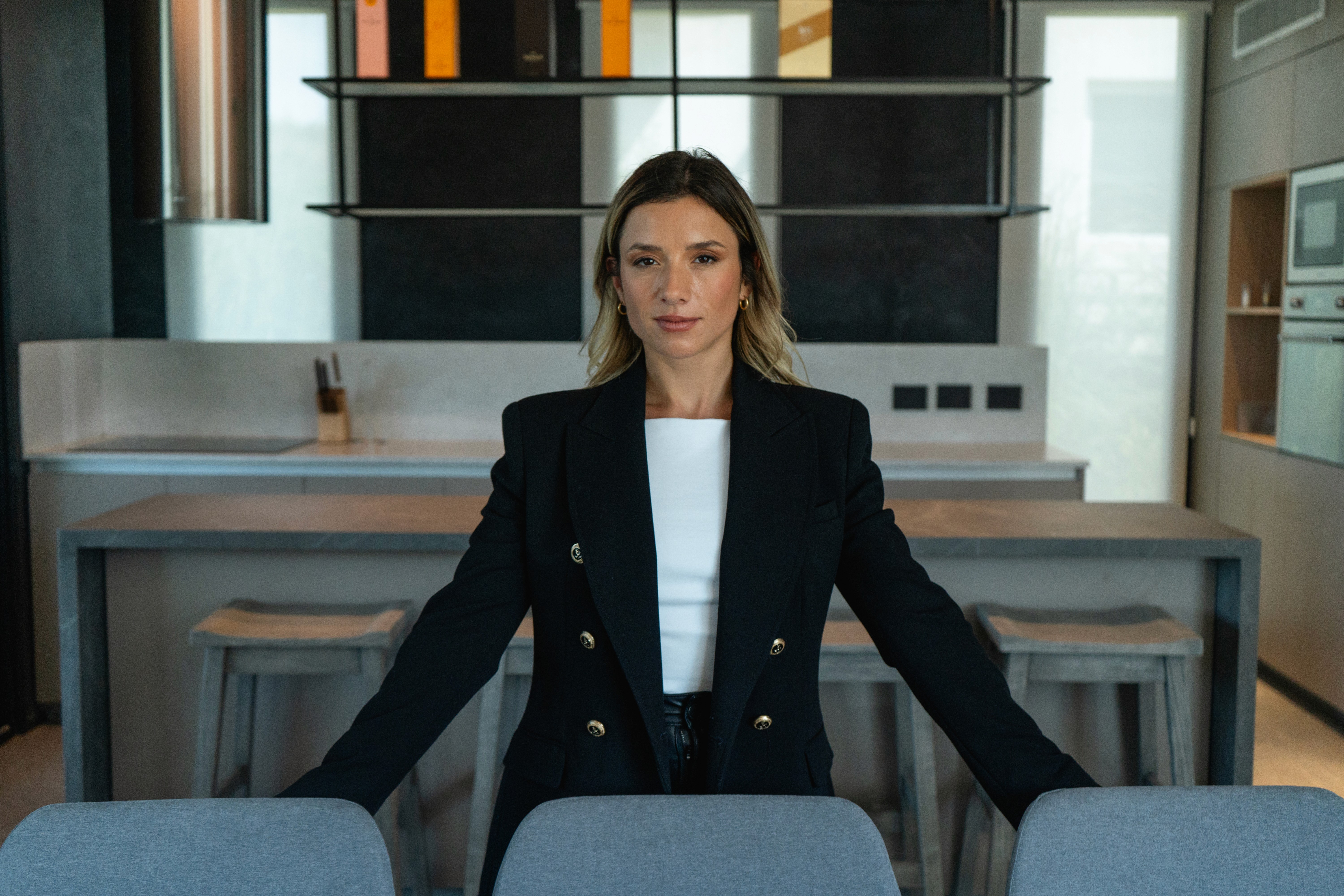 Mujer con blazer negro apoya ambas manos en sillas y mira de frente en cocina moderna minimalista.