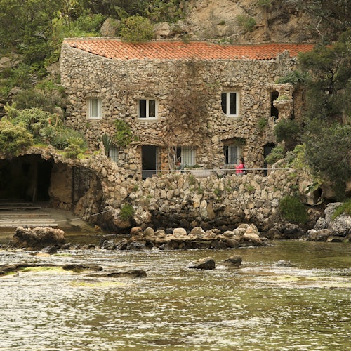 Maison en pierre avec toit rouge près de l'eau, entourée de verdure. Une personne en rose se tient sur un petit balcon.