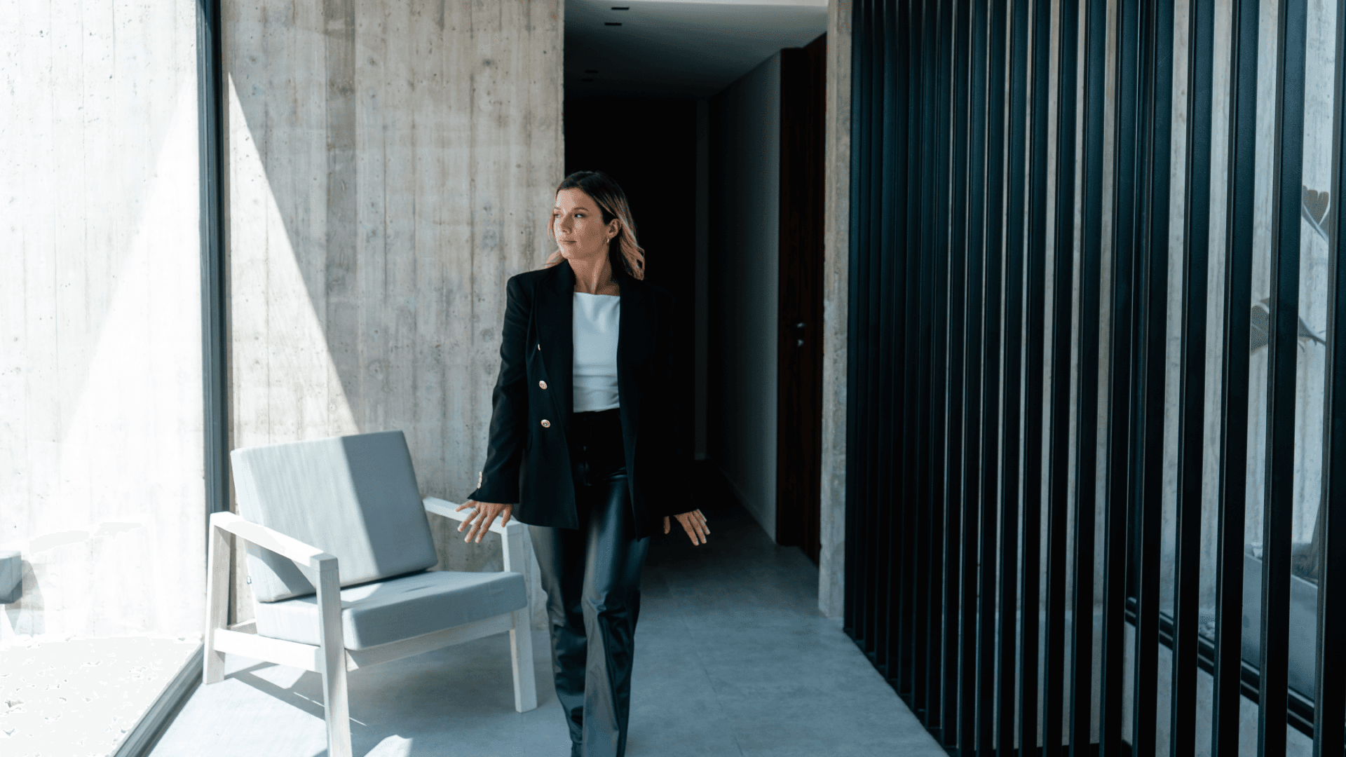 Mujer con blazer negro camina por pasillo moderno; pared de hormigón, paneles metálicos y butaca gris junto a ventanal.