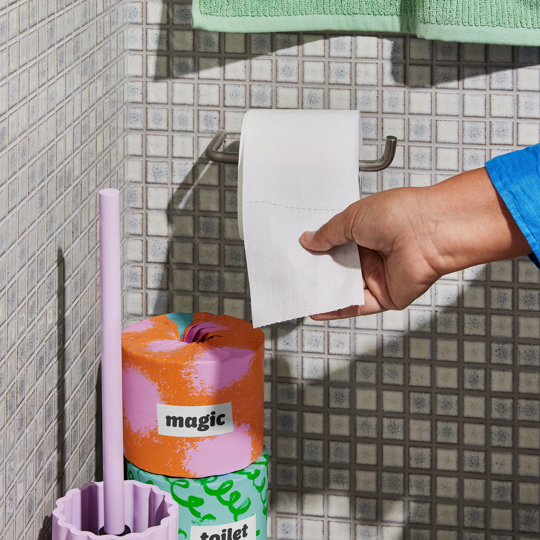 Hand pulling white toilet paper from a wall holder above a stack of colorful Who Gives A Crap rolls labeled magic and toilet