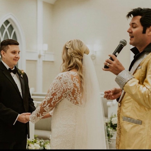 Ett par håller varandra i händerna under deras bröllopsceremoni medan en officiant, klädd i en guldjacka, talar i en mikrofon.