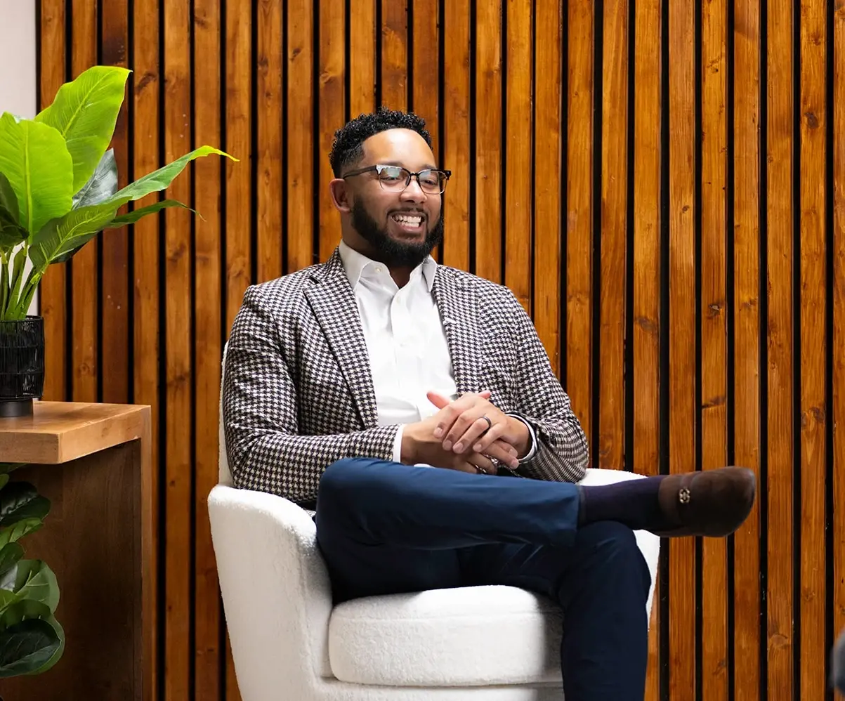 Chris Lewis sits in a modern white chair against a wooden wall, smiling during an interview or client meeting. The professional setting conveys warmth, expertise, and trust—reflecting the personal guidance offered by Chris Lewis Home Loans.