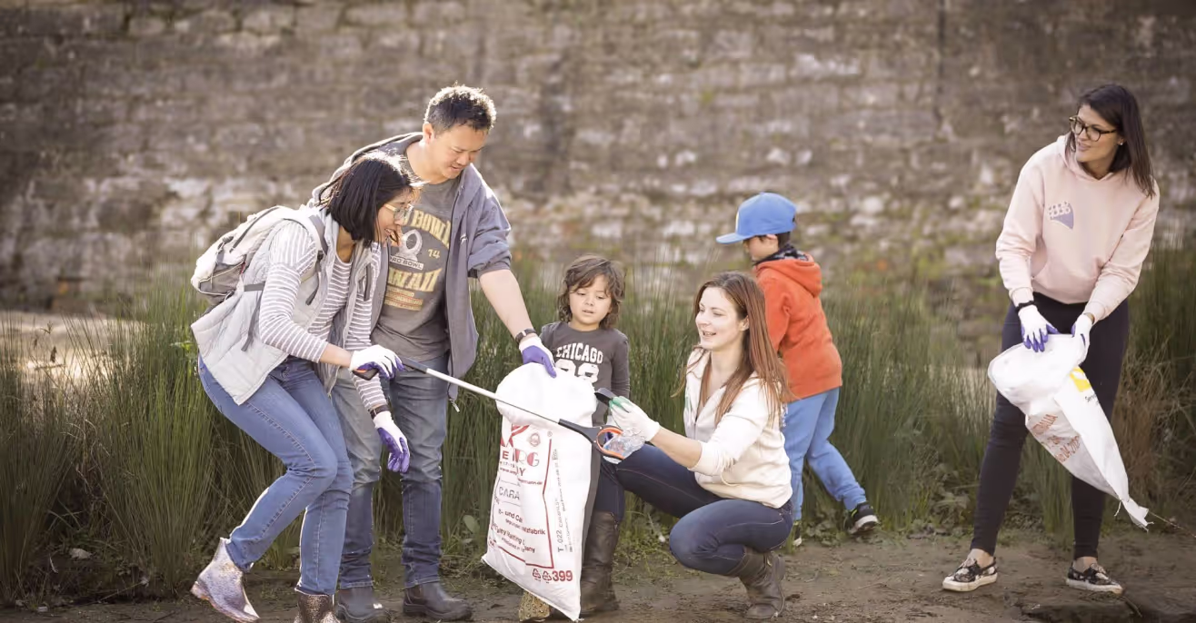A group of adults and children collecting litter beside a river using tongs and collection bags