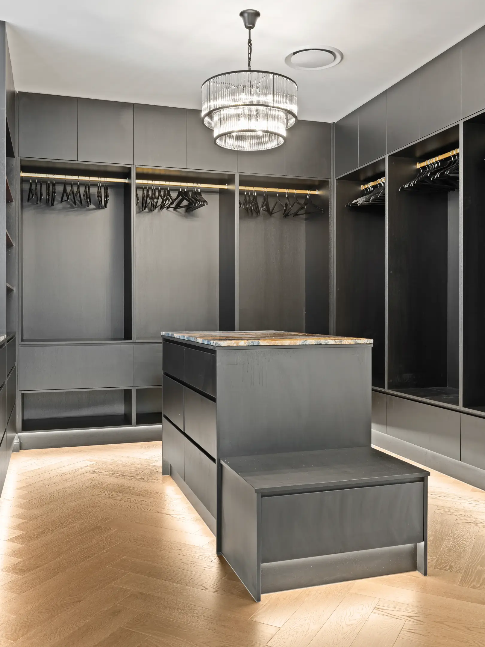 Luxurious custom walk-in wardrobe featuring matte black floor-to-ceiling cabinetry, a central island drawer unit with a stone top, timber herringbone floors, LED floor lighting, and a crystal chandelier.