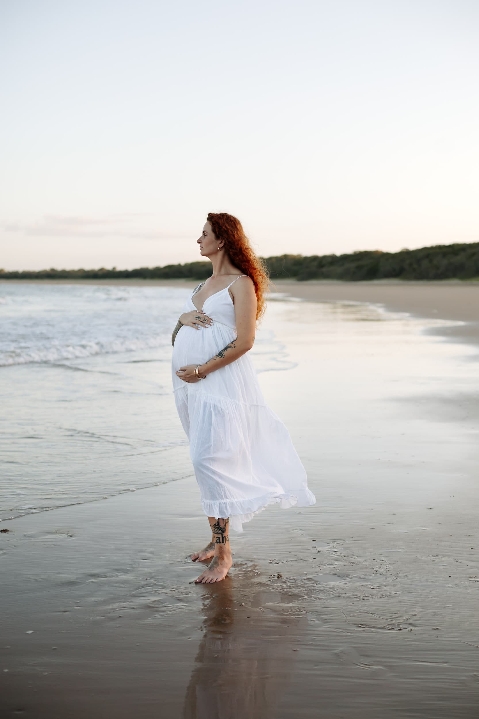 Expectant mother standing barefoot on the shoreline during a golden sunset maternity session on a Mackay beach.