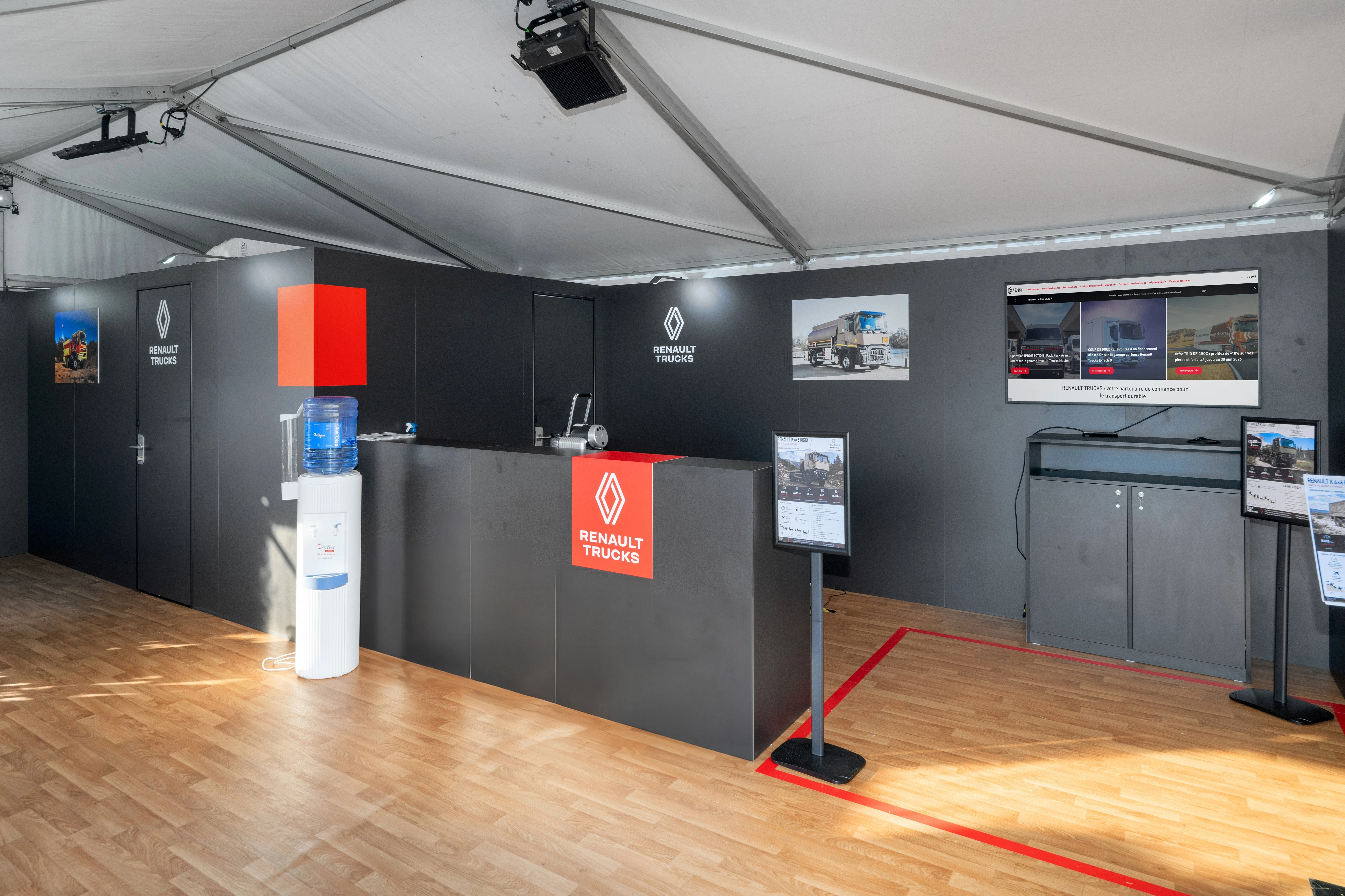 Stand extérieur Renault Trucks défense conçu par EXPACE à Eurosatory 2026 — camions militarisés 6x6 sable sur pelouse, façade anthracite et rouge, chapiteau d'accueil parquet