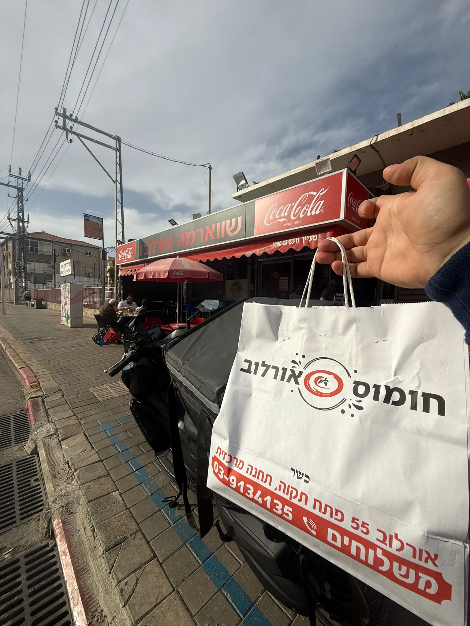 שקית חומוס מוחזקת מול מסעדה עם ישיבה בחוץ.