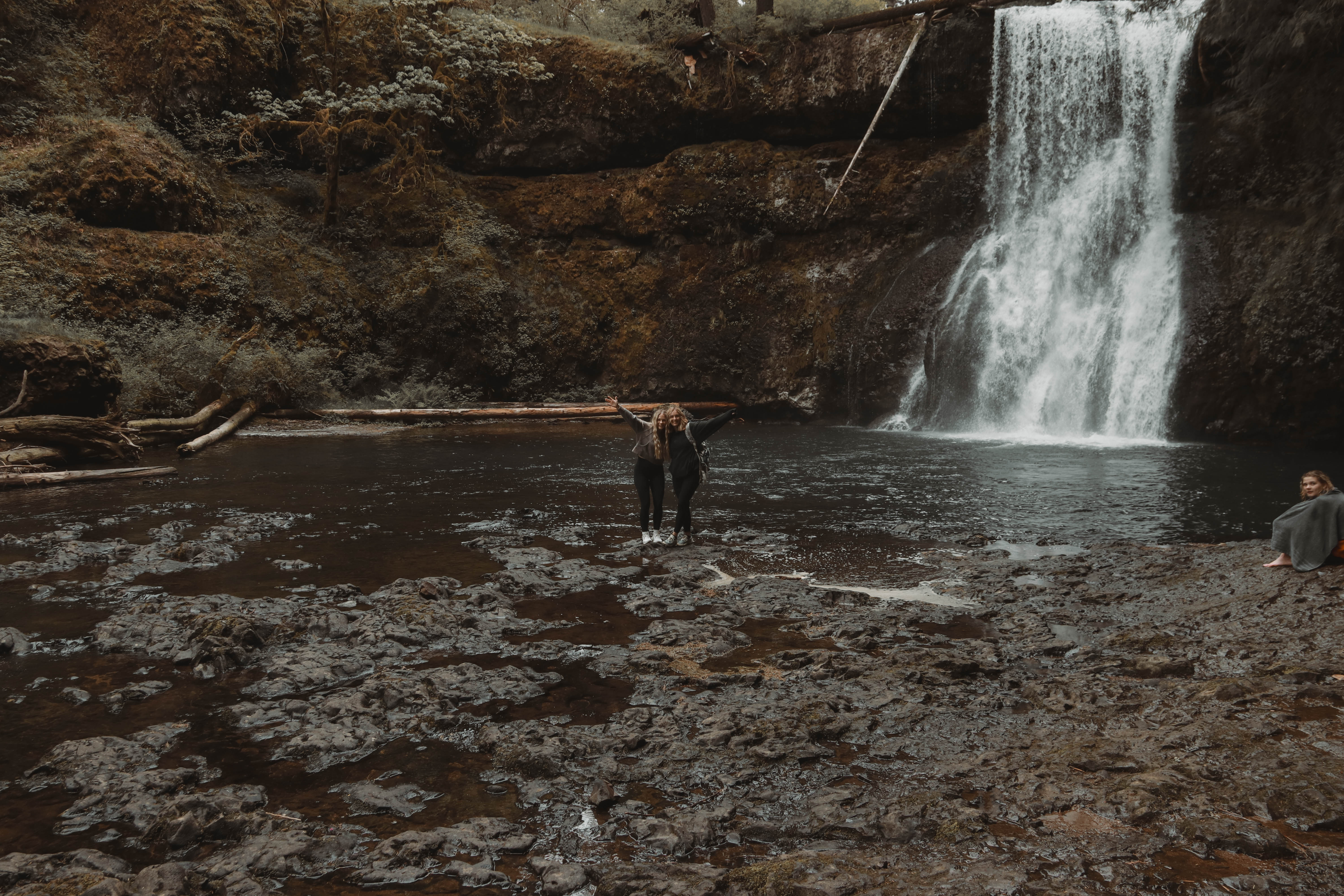 Two women holding their arms up in the air while standing by a waterfall in Oregon. 