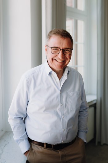 Älterer Männlicher Geschäftsführer im hellen Hemd steht locker vor einem Fenster im Büro von SLB.