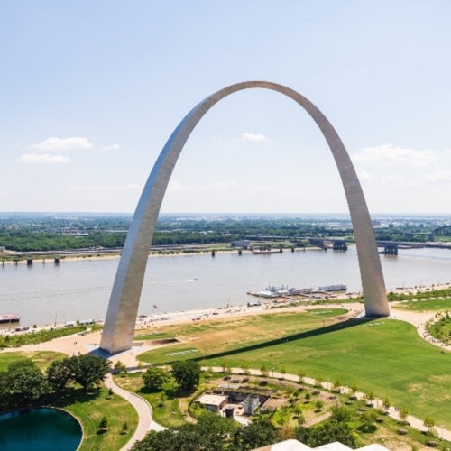 A large arch structure situated by a river with a bridge in the background, surrounded by green parks and pathways.