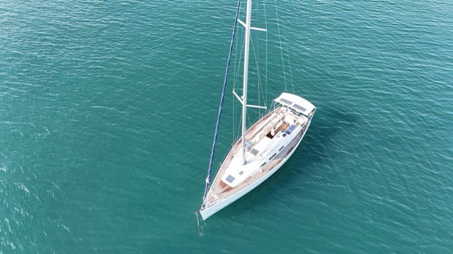 Overhead aerial top-down view of the Dufour 40 Performance sailing yacht at anchor in vivid turquoise Mediterranean water, showing the full deck plan including the teak decking, dark blue coachroof, solar panel, bimini, cockpit layout, and the elegant bow with blue carbon sprit against the clear shallow seabed below