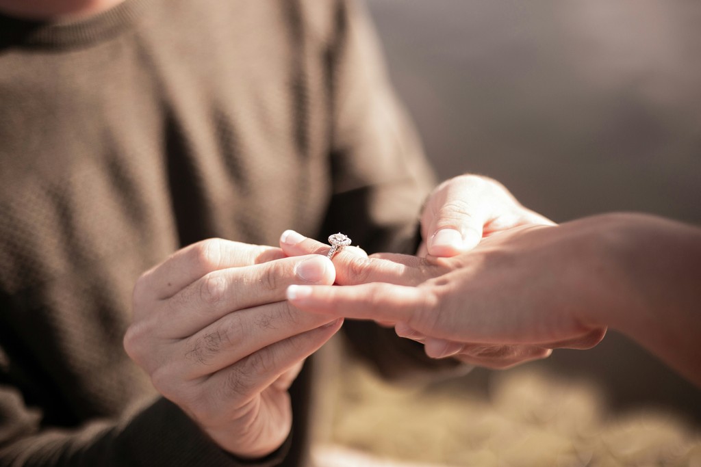Close-up of hands placing a diamond engagement ring on a finger with soft golden light in the background