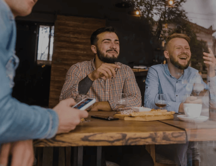 Un hombre está sentado en una mesa, sonriendo y sosteniendo una bebida en una mano. Parece estar disfrutando de un momento social, quizás compartiendo una comida o bebidas con amigos.